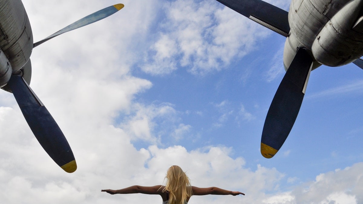 "A blonde woman standing with outstretched arms beneath the massive propellers of a Tu-95 strategic bomber in Keila, Estonia, symbolizing freedom and the vast scale of aviation history."